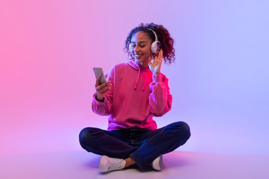 Relaxed black lady in pink hoodie sitting cross-legged, enjoying music with white headphones while browsing her smartphone in neon-lit background